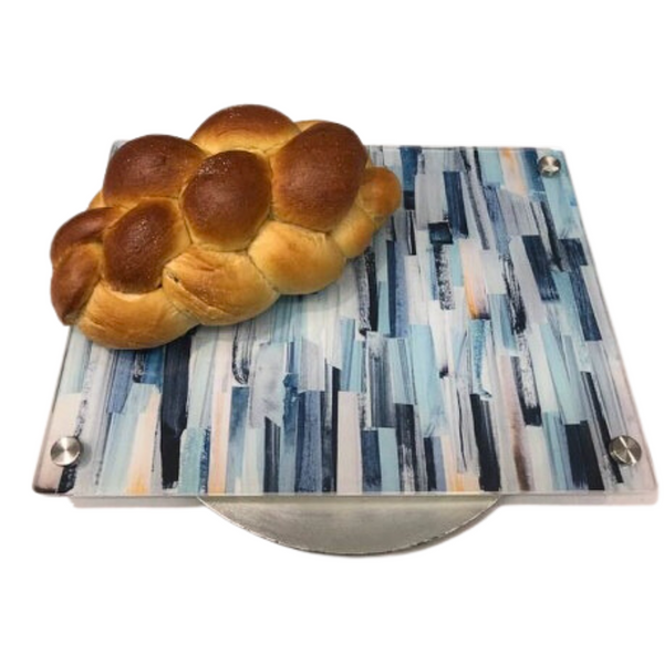 A loaf of challah bread sits on the Eye Candy Raised Serving Board Paint Stroke Blue, featuring blue and white brushstrokes on a square glass tray with a round metallic base and acrylic board.