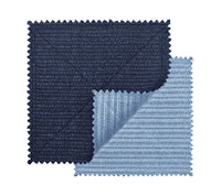 The Kim Seybert Vista Reversible Placemat s/4 in Navy & Periwinkle features two square, ribbed-textured placemats with zigzag edges; the navy piece overlaps the periwinkle one, whose corner is folded to show both reversible sides.