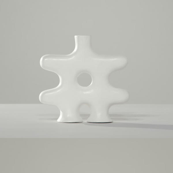 The Nedjma Vase by Schwung Design is a modern, abstract white ceramic piece with an irregular shape, round cutout center, and two short base supports. Handcrafted, it stands on a light surface against a plain gray background.