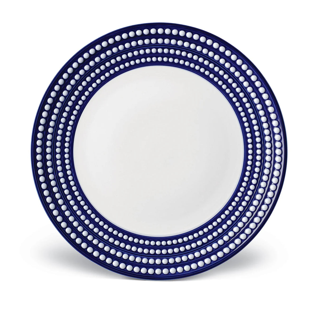 The L'Objet Perlèe Blue Dinnerware features a round ceramic plate with a white center and wide dark blue rim, accented by rows of evenly spaced white dots—ideal for fans of blue and white tableware.