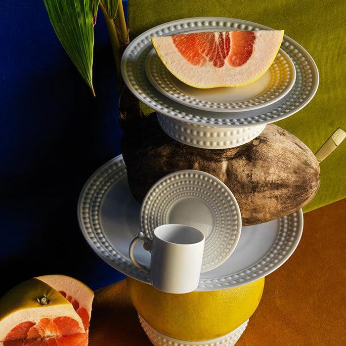 A creative arrangement of L'Objet Perlèe White Dinnerware, a teacup, grapefruit slice, yam, and green leaf set against a vibrant blue, green, and brown background.