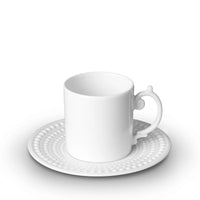 A white ceramic teacup with an ornate handle rests on a matching saucer from L'Objet's Perlèe White Dinnerware, both featuring a raised dotted pattern, displayed on a white background.