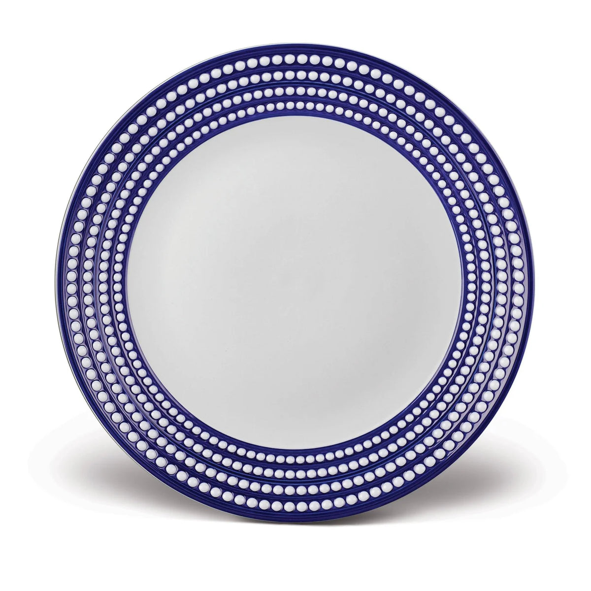 The Perlèe Blue Dinnerware by L'Objet features a round white plate with a blue rim and concentric rows of white dots, exemplifying elegant blue and white design in fine Limoges porcelain.