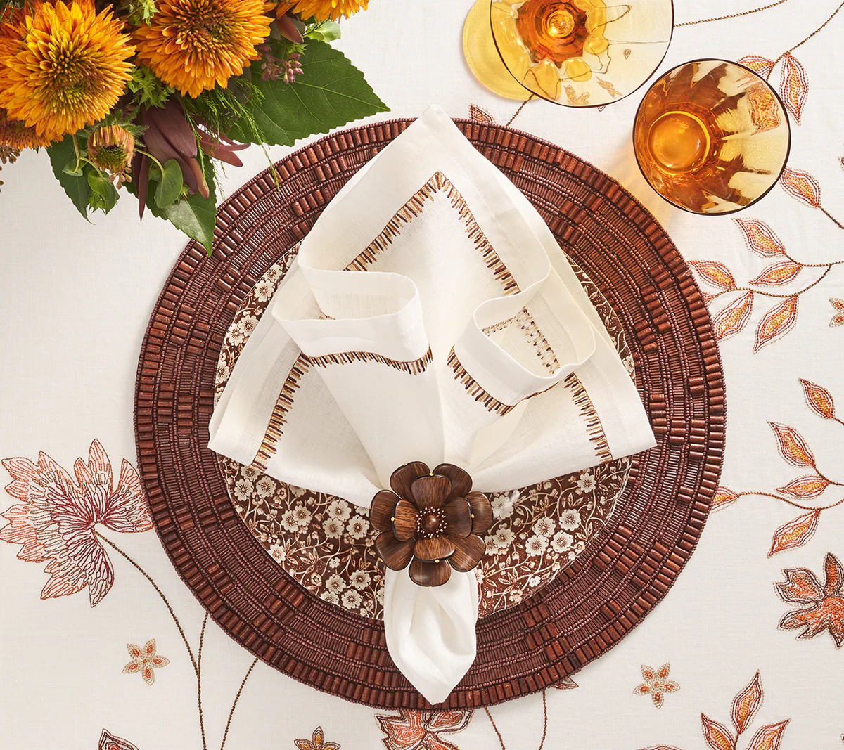 A fall-themed table setup featuring a white napkin in a brown flower-shaped ring on a Kim Seybert Wood Matrix Placemat, surrounded by autumn flowers and amber drinks atop a floral tablecloth.