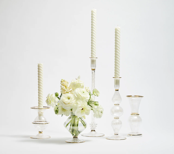 A white bouquet of ranunculus and hyacinth rests in the Kim Seybert Clear Rib Gold Detail Trumpet Vase, displayed beside three glass candlesticks with white twisted candles.