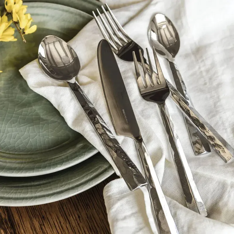 The Simon Pearce Charlotte Flatware 5 Piece Setting, with hand-forged hammered stainless steel handles, rests on a white napkin beside stacked green plates on a wooden table. Yellow flowers peek in from the corner.