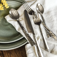 The Simon Pearce Charlotte Flatware 5 Piece Setting, with hand-forged hammered stainless steel handles, rests on a white napkin beside stacked green plates on a wooden table. Yellow flowers peek in from the corner.