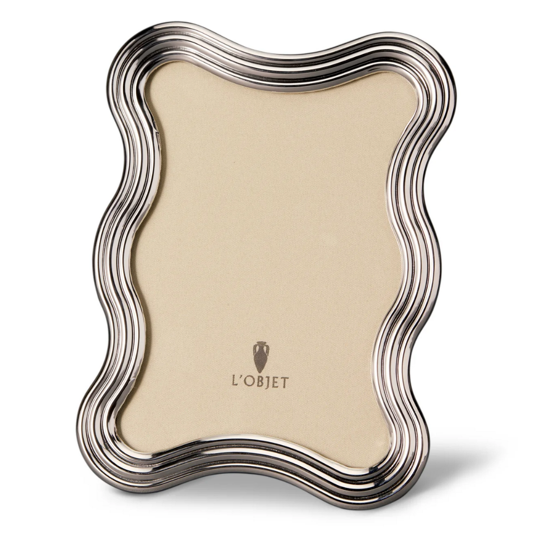 The L'Objet Ripple Frame Platinum features wavy, rippled silver edges with a platinum finish and a beige backing displaying the L'Objet logo. This modern frame stands upright on any flat surface.