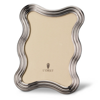 The L'Objet Ripple Frame Platinum features wavy, rippled silver edges with a platinum finish and a beige backing displaying the L'Objet logo. This modern frame stands upright on any flat surface.