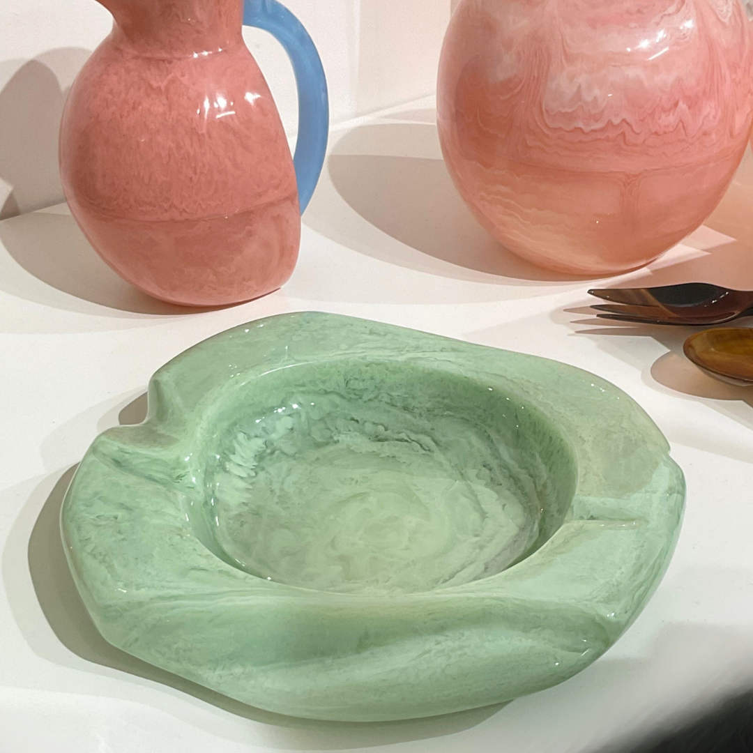 The Malibu Resin Dish by Lily Juliet, featuring wavy edges and a light green hue, rests on a white surface with two pink and blue marbled glass pitchers and several spoons in the background.