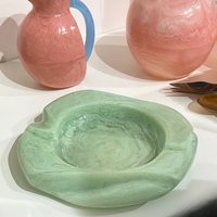 The Malibu Resin Dish by Lily Juliet, featuring wavy edges and a light green hue, rests on a white surface with two pink and blue marbled glass pitchers and several spoons in the background.