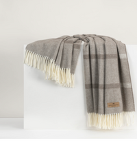 The Montauk Stripe Throw by Lands Downunder, featuring a soft gray herringbone plaid and fringed cream edges, is draped over a white block against a neutral background. This reversible throw is expertly crafted in Italy.