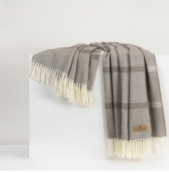 The Montauk Stripe Throw by Lands Downunder, featuring a soft gray herringbone plaid and fringed cream edges, is draped over a white block against a neutral background. This reversible throw is expertly crafted in Italy.