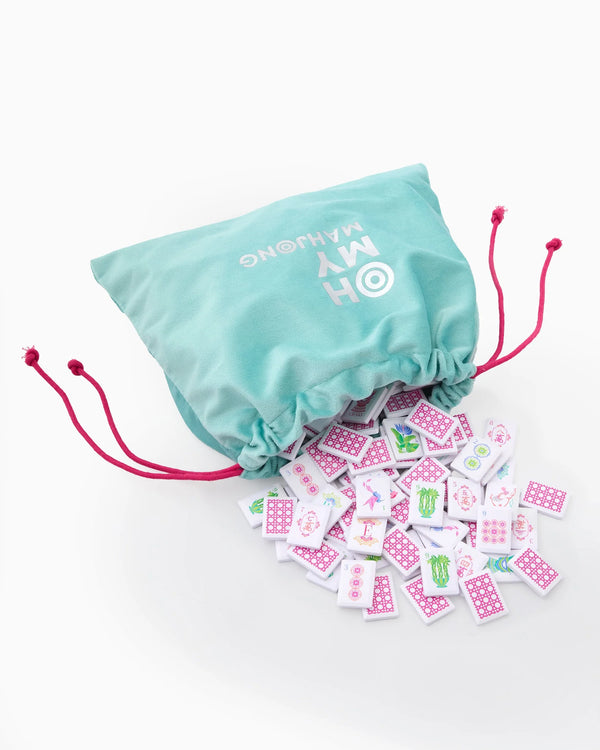 The Mahjong Debutante Travel Tile Set by Oh My Mahjong features a teal drawstring bag with pink cords, revealing colorful patterned tiles—perfect for introducing mahjong to kids anywhere.