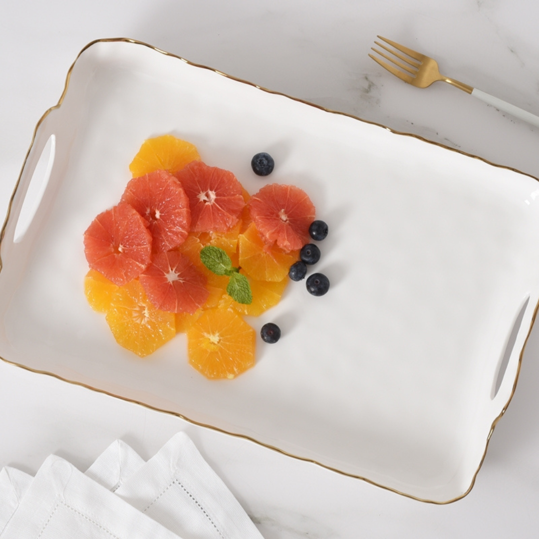 The PAMPA BAY Portofino Handle Rectangle Tray with a gold border displays sliced oranges, grapefruit, blueberries, and mint. Nearby on the marble surface are a gold fork and folded white napkins.