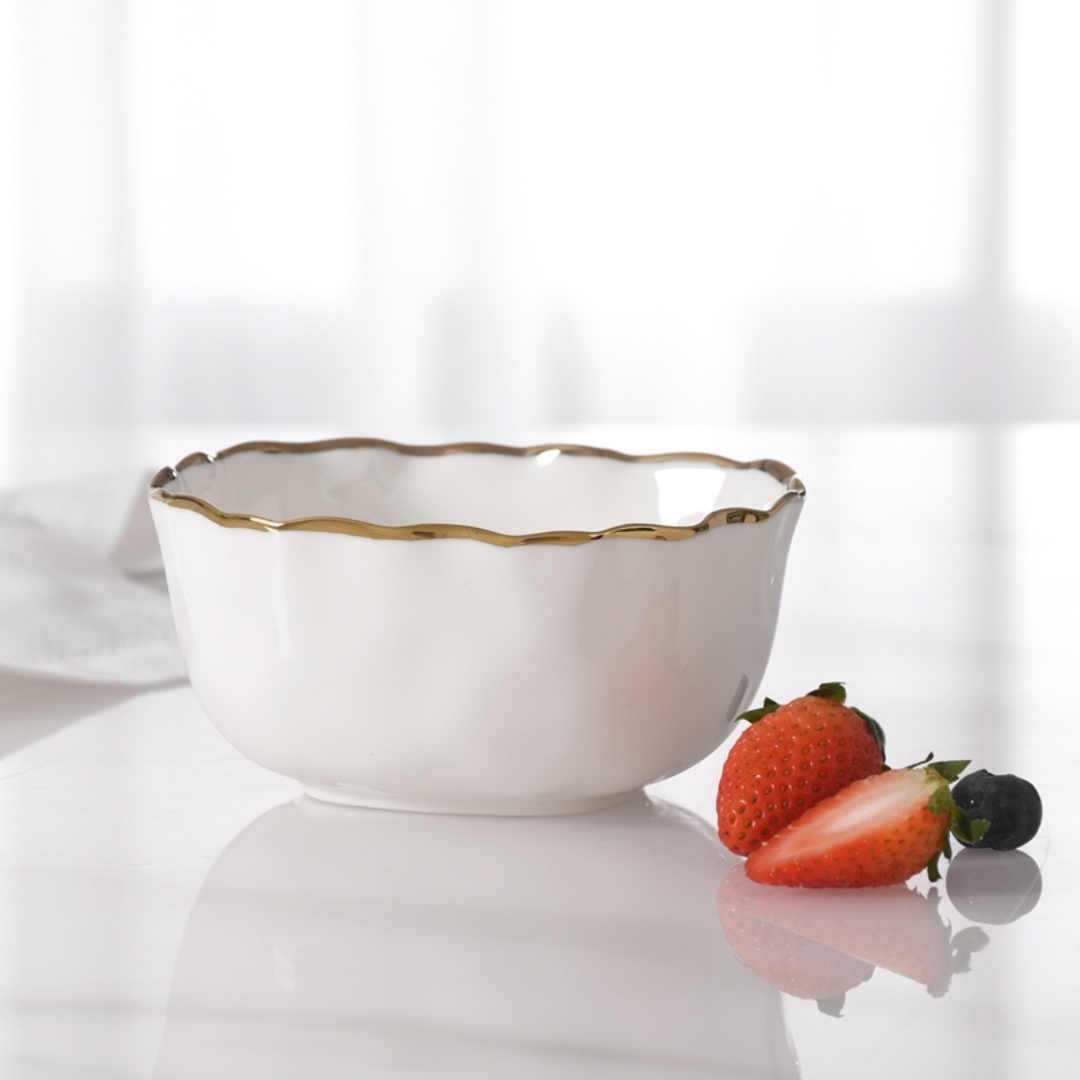 The PAMPA BAY Portofino Snack Bowl Gold with elegant gold trim rests on a glossy surface beside a whole strawberry, a halved strawberry, and a blueberry. A white cloth is visible in the background.