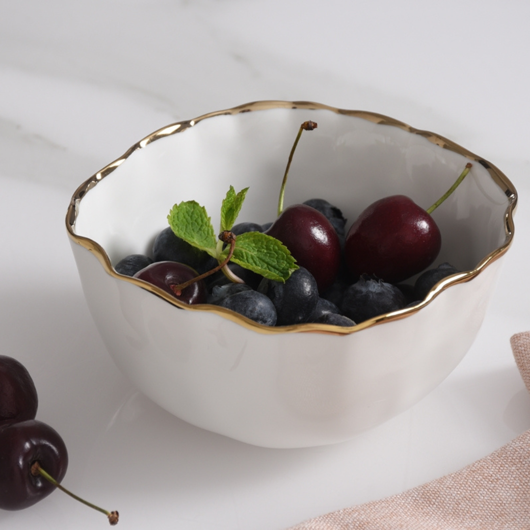The PAMPA BAY Portofino Snack Bowl Gold, filled with blueberries, cherries, and mint, rests on a white surface beside a few cherries and a pink napkin—ideal for oven to table serving.
