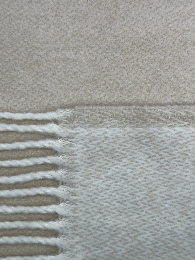 A close-up of A Soft Idea's Double Faced Solid Mini Chevron Throw shows its textured beige and cream weave, with fringe on the left and a smooth solid edge on the right, highlighting its reversible design.