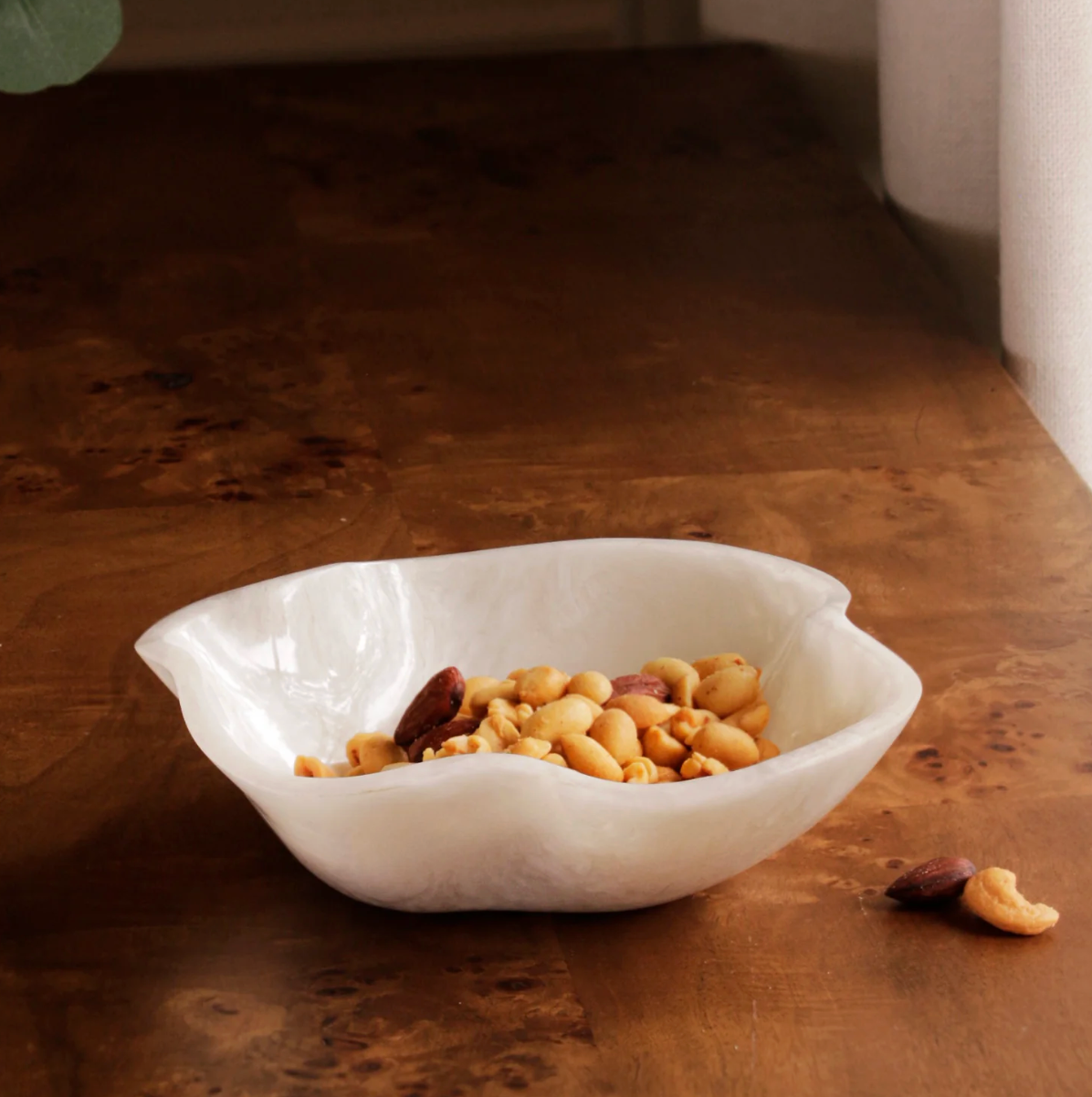 The Beatriz Ball Resin Alabaster Mini Bowl, with its wavy white edges, holds mixed nuts and sits on a wooden table near a window draped with a light-colored curtain.