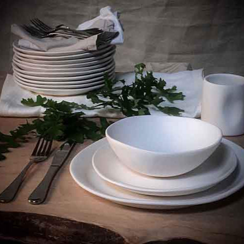 A rustic table set with Alex Marshall Studios Slim Dinnerware Matte White—stacked plates, bowl, and cup—all microwave and dishwasher safe—paired with cutlery on a napkin and sprigs of greenery on a wooden surface.