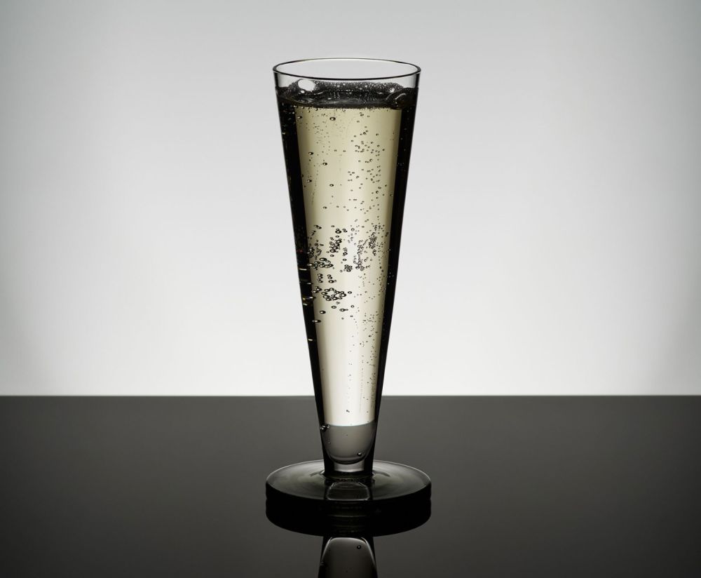 A Tom Dixon Puck Flute Glass from the Set of 2, filled with sparkling wine and rising bubbles, stands on a reflective black surface against a minimalist gray background.