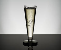 A Tom Dixon Puck Flute Glass from the Set of 2, filled with sparkling wine and rising bubbles, stands on a reflective black surface against a minimalist gray background.