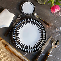 A table setting featuring Molecot's Punk Dinnerware—black and white geometric plates—on gray marble, with dark-handled cutlery, a white napkin, a glass, and a vase holding a pink tulip.