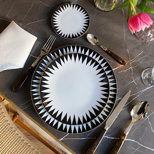 A table setting featuring Molecot's Punk Dinnerware—black and white geometric plates—on gray marble, with dark-handled cutlery, a white napkin, a glass, and a vase holding a pink tulip.