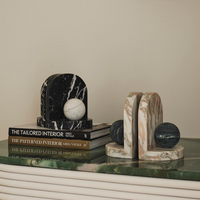 Two sets of Greg Natale's Blitz Marble Bookend Set—one black and white, one beige with green—enhance stacked design books on a green marble-topped table, creating an elegant ambiance in this luxury home decor.