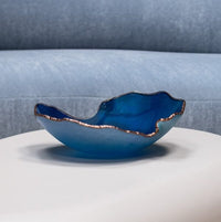 A wavy, dark blue Mapa Azul With Bronze Trim Bowl by Gardeco sits on a white surface, with a light blue sofa in the background.
