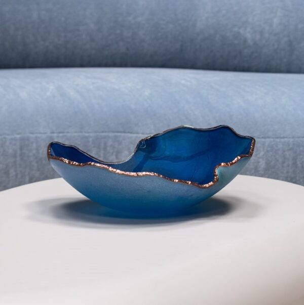 A wavy, dark blue Mapa Azul With Bronze Trim Bowl by Gardeco sits on a white surface, with a light blue sofa in the background.