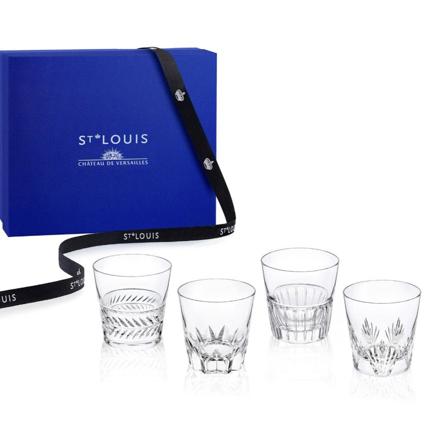 A blue Saint-Louis/Hermes Château de Versailles gift box with a black ribbon stands behind four clear Kings Hall Tumblers, each intricately cut in a unique design.