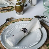 A white napkin rolled inside a Christofle Babylon Silver Plated Napkin Ring Set 4 with a braided design, resting on a white dinner plate.