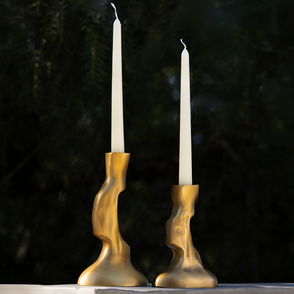 Two off-white taper candles are displayed in Frankie Taper Candle Holder Brass - by Dumae, placed side by side against a dark, blurred foliage background.