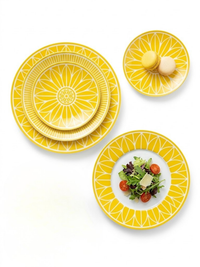 Christofle’s Malmaison Riviera Dinnerware, featuring yellow and white patterns, is displayed on a white background—one plate with salad, another with macarons—evoking the elegance of French Riviera dining.