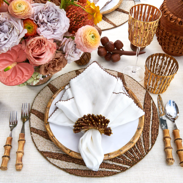 A table setting with a floral centerpiece, woven placemat, bamboo-handled cutlery, Kim Seybert Bamboo Glassware Gold Set of 4, and a white napkin with a brown beaded ring on a white plate creates an elegant, earthy tablescape.