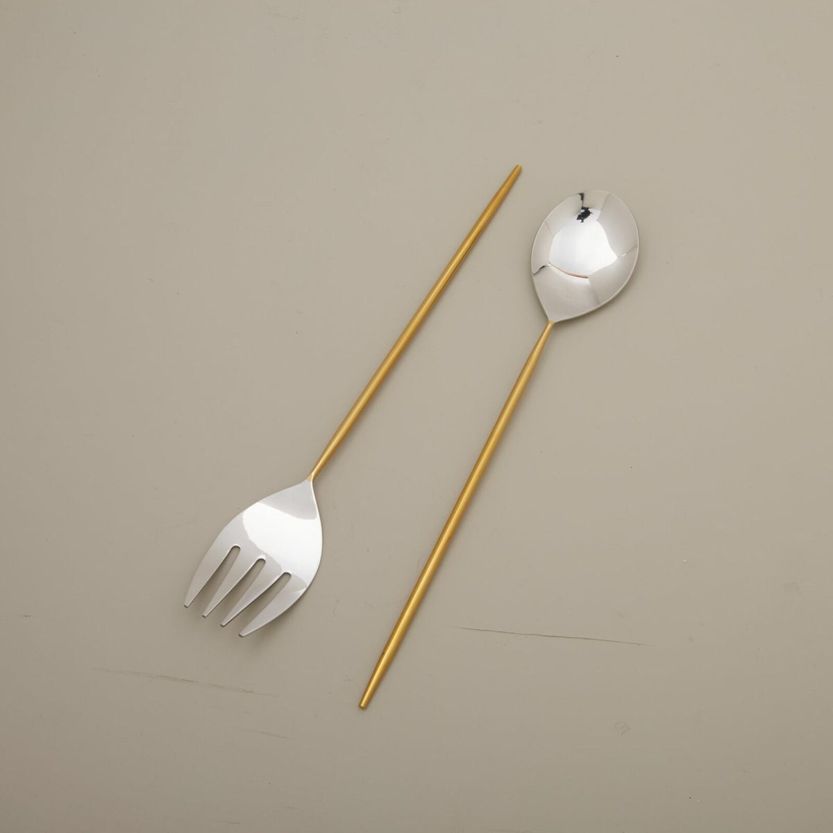 The Be Home Mixed Silver & Gold Serving Set, featuring a sleek fork with gold-colored handle on the left and a matching spoon on the right, rests on a plain light beige surface.