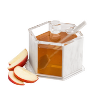 A Lucite Hexagon Apple Honey Jar with Spoon in White Marble by BT- Shalom, filled with honey, is displayed next to three slices of red-skinned apple on a white background—an elegant touch of holiday table decor perfect for Rosh Hashanah celebrations.