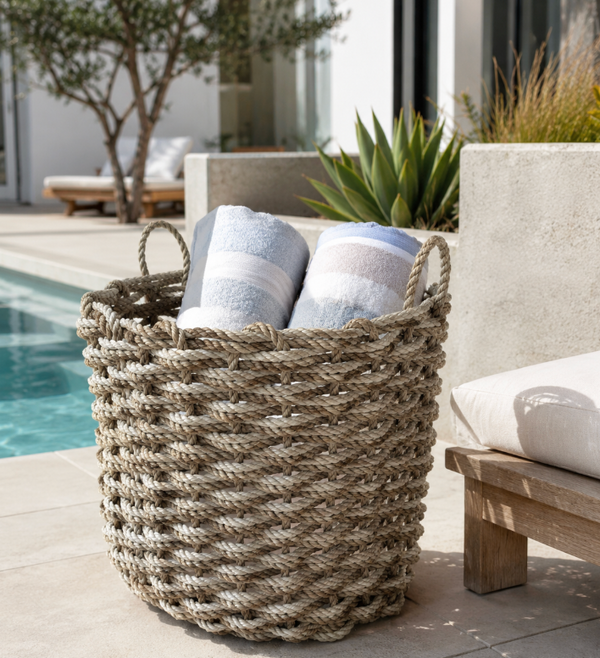 The Rope Co’s Coastal Outdoor Basket in Sand & Oyster, filled with two rolled striped towels, sits poolside on a patio near a lounge chair, surrounded by greenery and framed by a modern building.