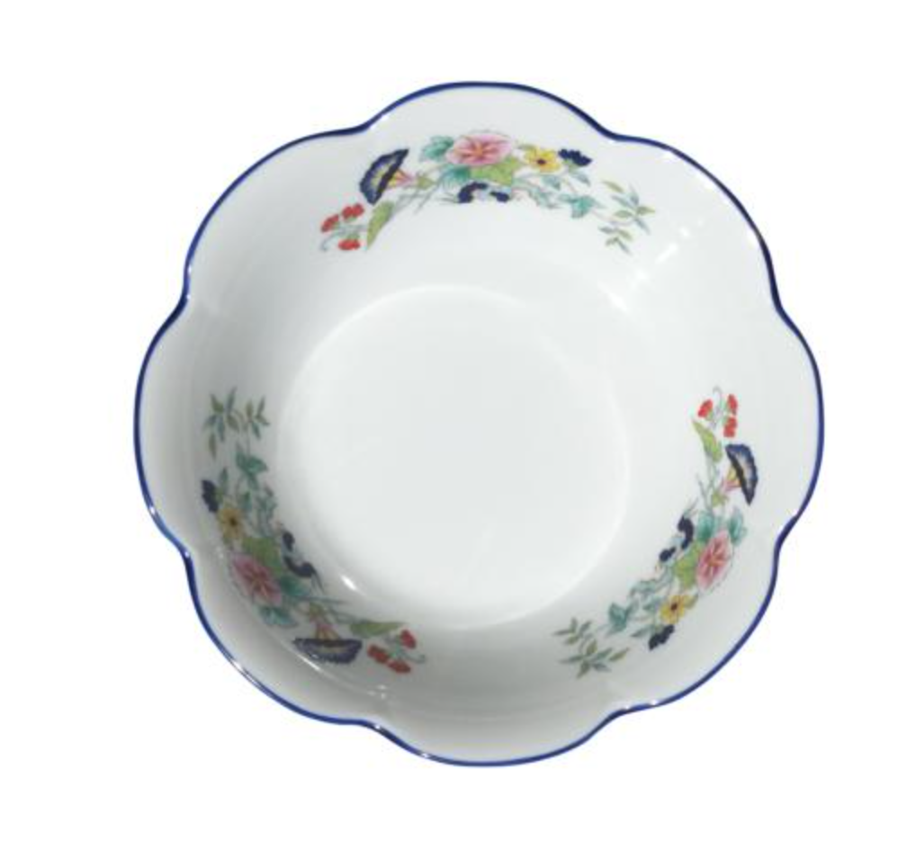 A white porcelain bowl with a scalloped edge and blue rim from Royal Limoges’ Nymphea Paradis Bleu collection, featuring colorful floral patterns and green leaves on the inner sides.