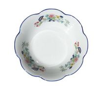 A white porcelain bowl with a scalloped edge and blue rim from Royal Limoges’ Nymphea Paradis Bleu collection, featuring colorful floral patterns and green leaves on the inner sides.