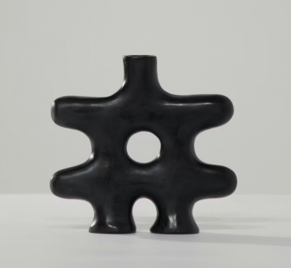 The Nedjma Vase - Black by Schwung Design is a modern black abstract ceramic vessel featuring a central hole and four rounded protrusions, evoking sculptural curves or an organic gear-like form. Crafted with artistry inspired by handmade Mediterranean techniques, it sits against a light gray background.