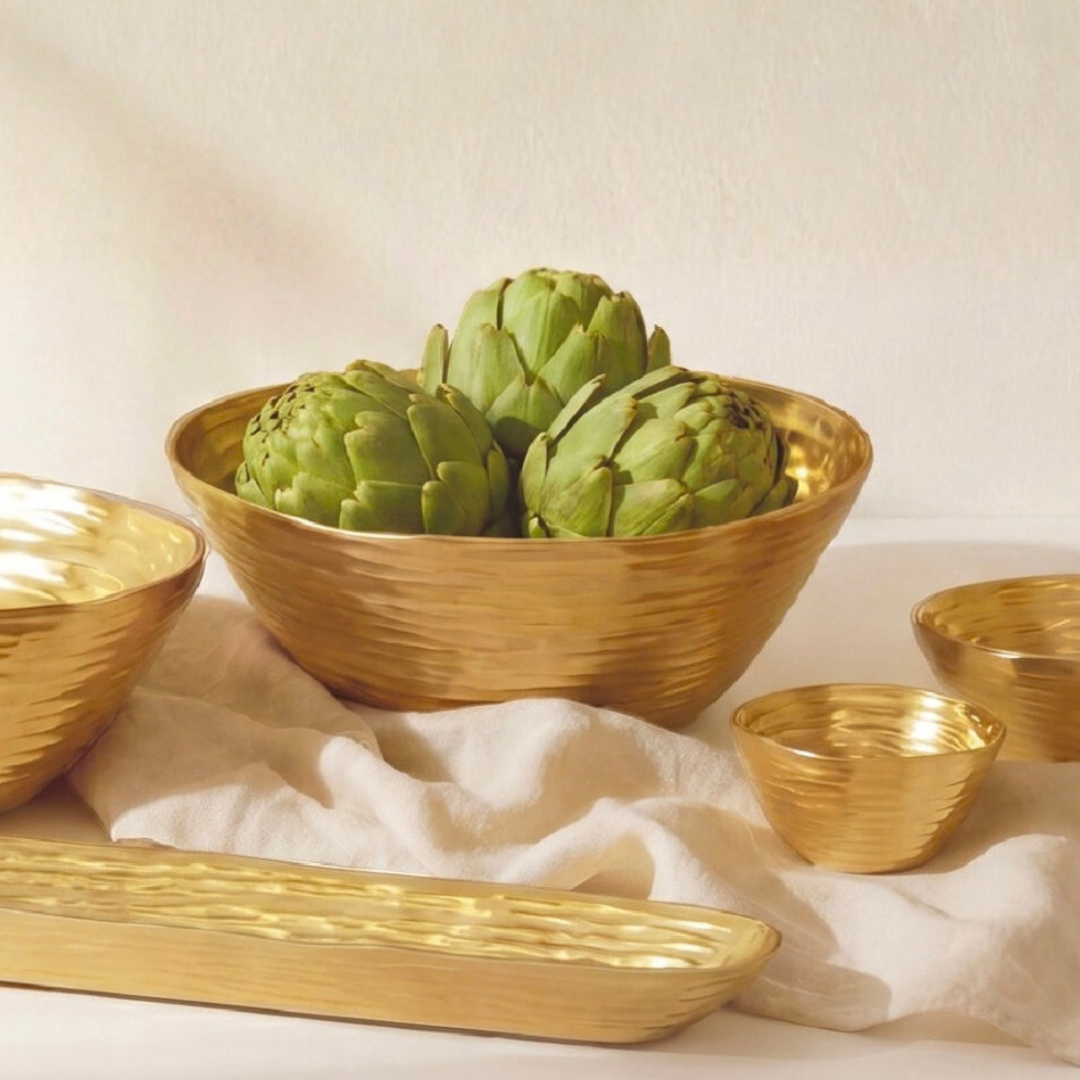 The Dorrato Gold Bowl - by PAMPA BAY is arranged on a cream cloth with trays and green artichokes, creating a warm, elegant, and minimalistic setting.