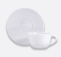 A white ceramic teacup with handle and matching saucer, both featuring subtle circular patterns, are displayed on a light gray background—an elegant piece from Bernardaud’s Origine Dinnerware White collection.