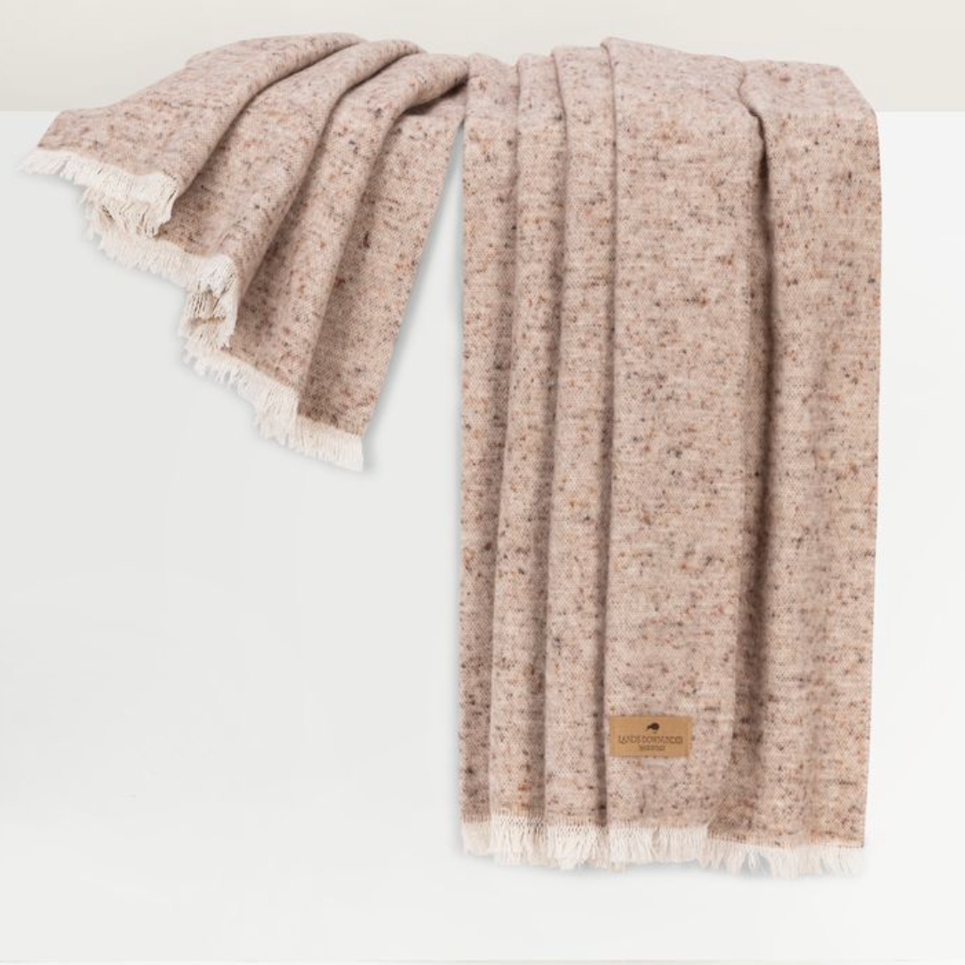 A soft, speckled beige Cinder Mist Throw by Lands Downunder, crafted from wool and finished with white fringe edges, is draped over a white surface. This modern throw features a small leather tag sewn on one corner, adding a touch of understated elegance.