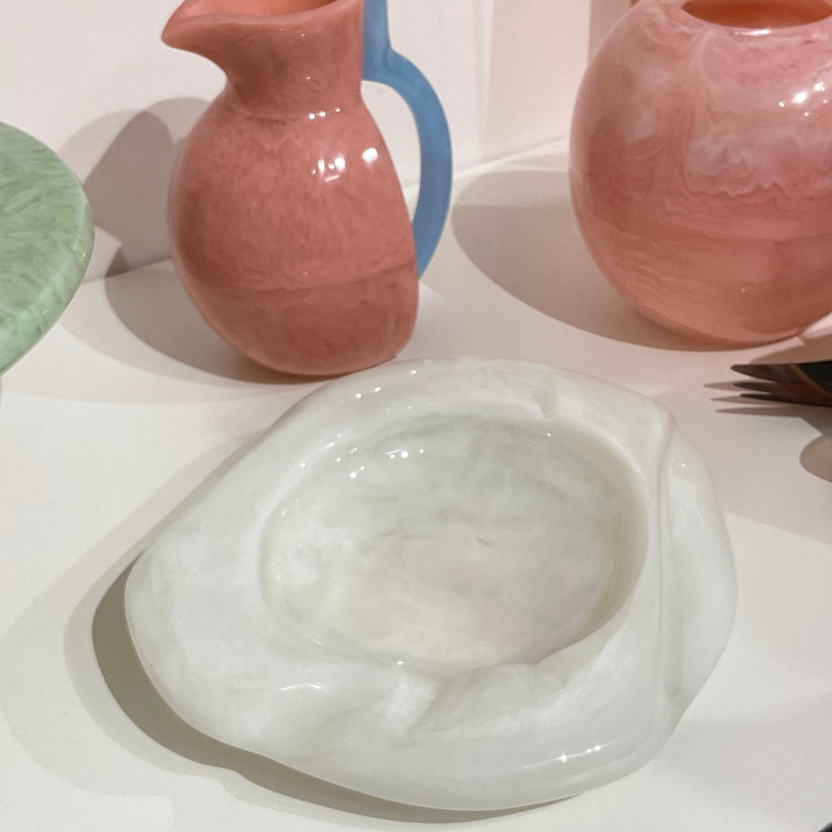 A white Malibu Resin Dish by Lily Juliet sits on a table beside two pink vases—one with a blue handle—and part of a green plate, all arranged in a soft pastel palette that accentuates the dish’s unique shape.