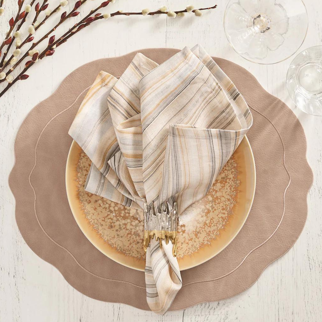 A place setting featuring the Kim Seybert Tailored Placemat Taupe, a beige and gold-rimmed plate, striped linen napkin in a gold/silver ring, on a white wooden table with pussy willow branches and glassware.