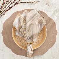 A place setting featuring the Kim Seybert Tailored Placemat Taupe, a beige and gold-rimmed plate, striped linen napkin in a gold/silver ring, on a white wooden table with pussy willow branches and glassware.