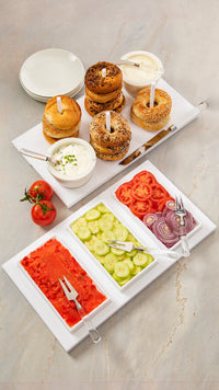 The Inspired Generations Bright White Brunch Board is stylishly arranged with bagels, cream cheese, and platters of sliced tomatoes, cucumbers, red onions, and smoked salmon for an organized brunch display. Plates and utensils are also included, making this serving board a perfect choice for elegant entertaining.