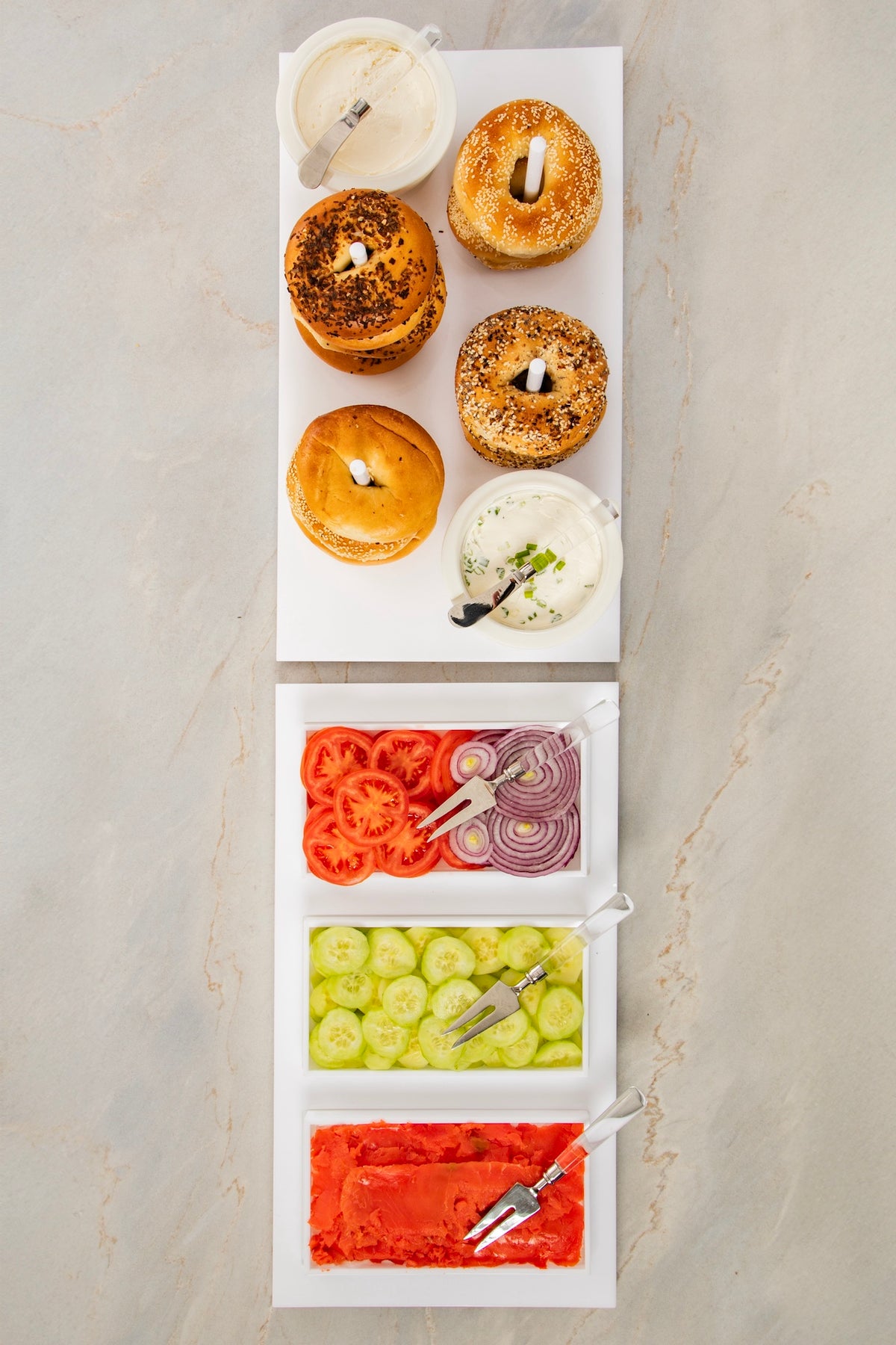Four bagels with spreads are elegantly arranged on the Inspired Generations Acrylic Bagels On Board Tray in White, accompanied by three trays of sliced tomatoes, red onions, cucumbers, and smoked salmon—each with convenient serving utensils—all set atop a light marble surface to create the perfect brunch board.